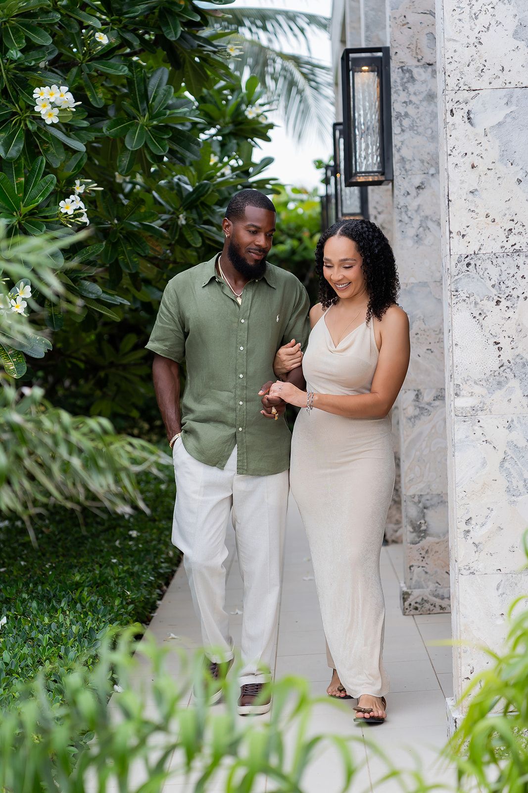 A couple walks together along a garden path, man in green shirt and white pants, woman in beige dress.