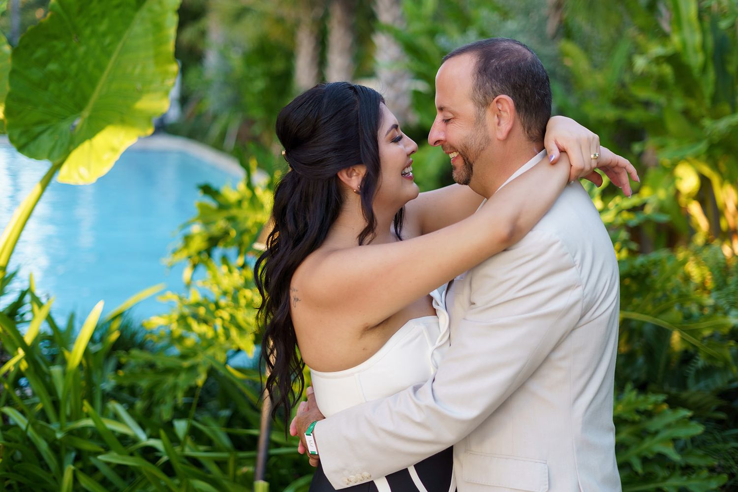 A couple embraces and smiles in a lush tropical garden with a pool in the background.