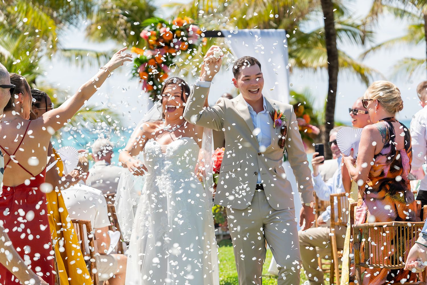 Joyful newlyweds walk through confetti shower at outdoor tropical beach wedding ceremony with palm trees.