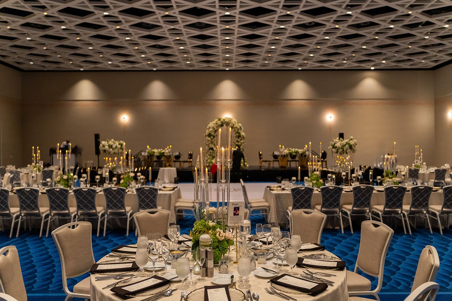 Elegant ballroom wedding reception with blue carpet, round tables, floral centerpieces, and tall gold candelabras under a coffered ceiling.