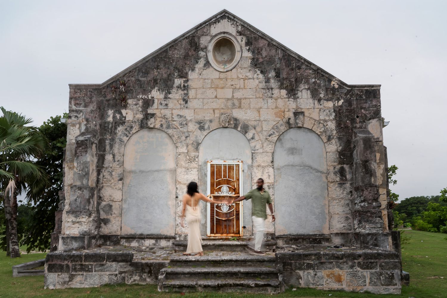 Couple standing before ancient stone church ruins with arched windows in a tropical setting.