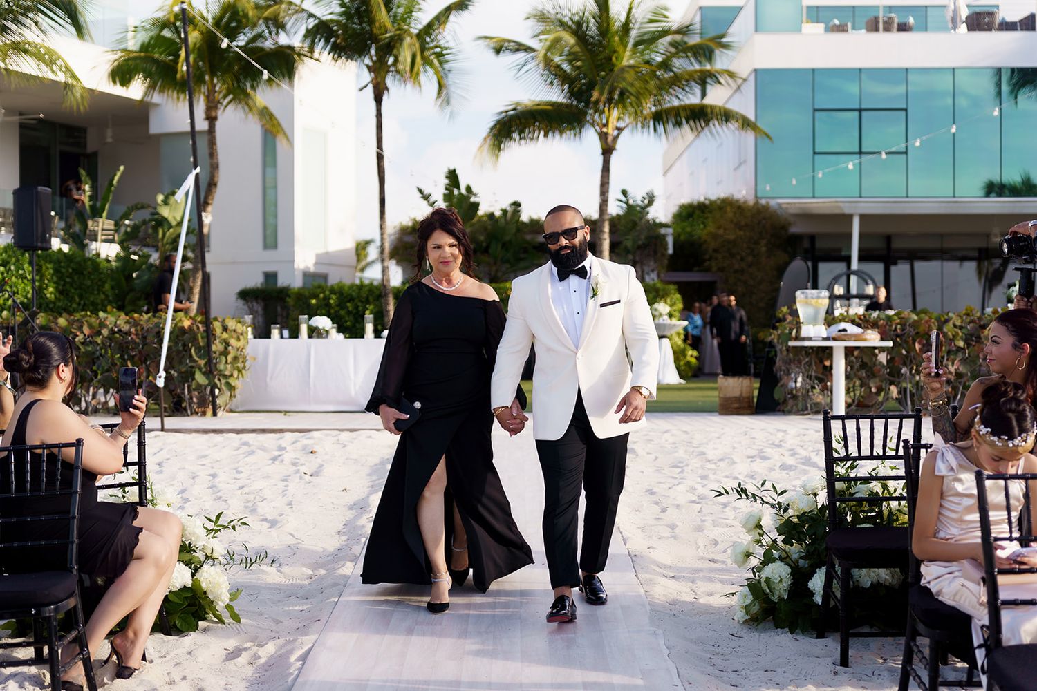 Couple in formal attire walking hand-in-hand at an elegant outdoor beach wedding ceremony with palm trees.