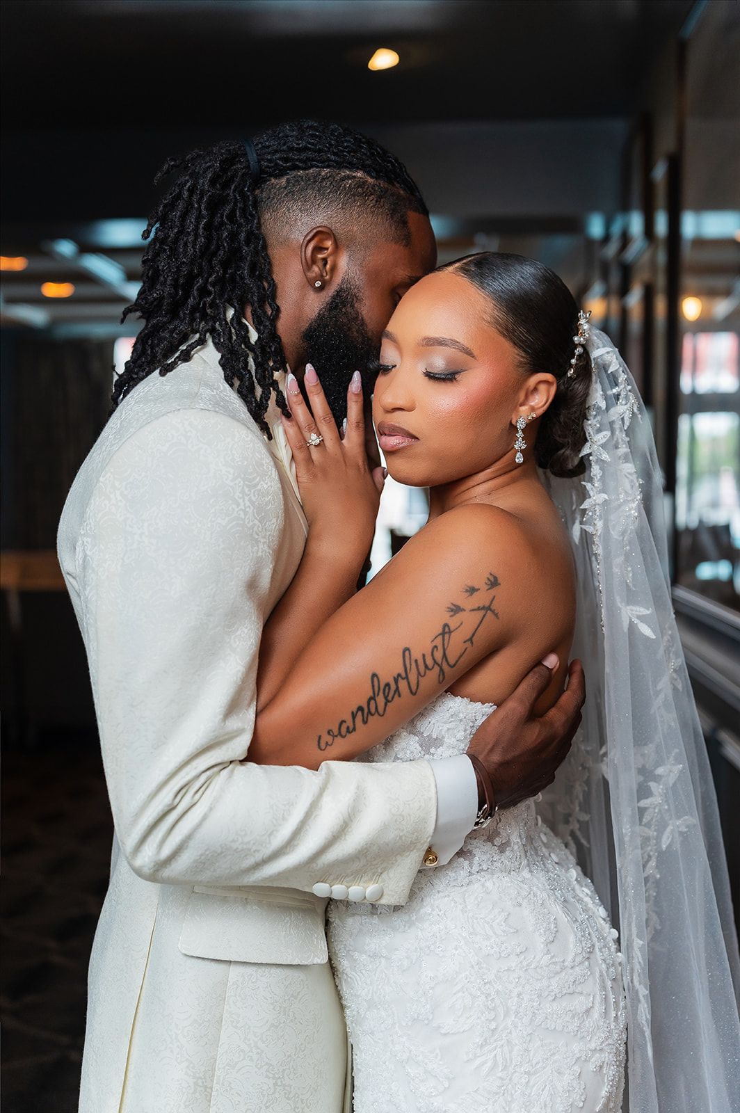 A bride and groom share an intimate moment in wedding attire, she in a lace gown with veil, he in a cream suit.