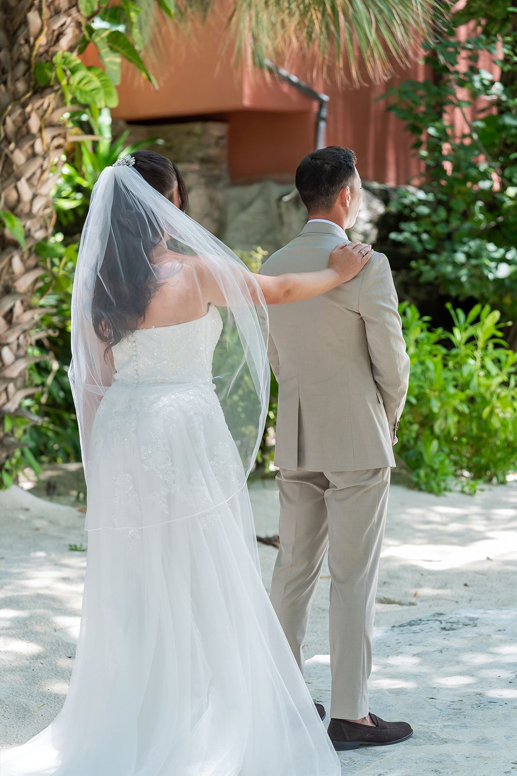Bride in white gown and veil touches groom's shoulder in beige suit during outdoor wedding first look.