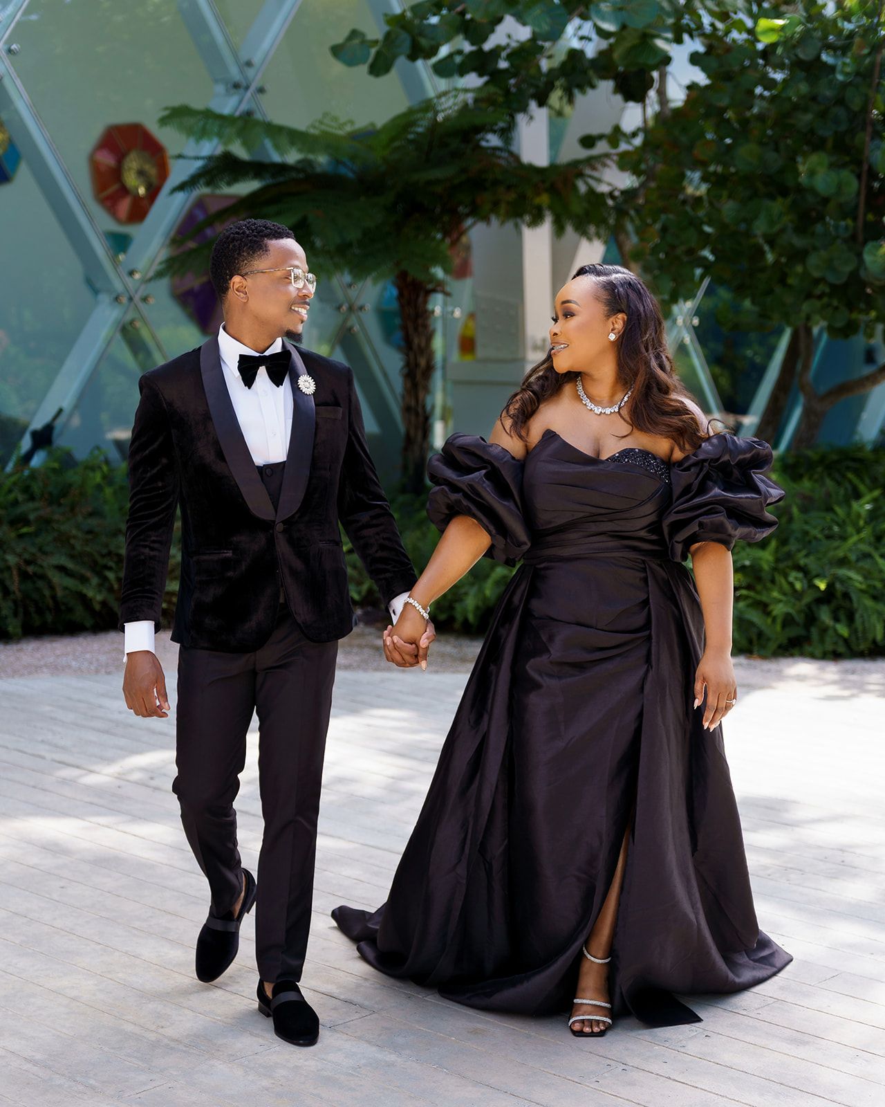 Elegant couple in matching black formal attire holding hands and walking together at an outdoor event venue.