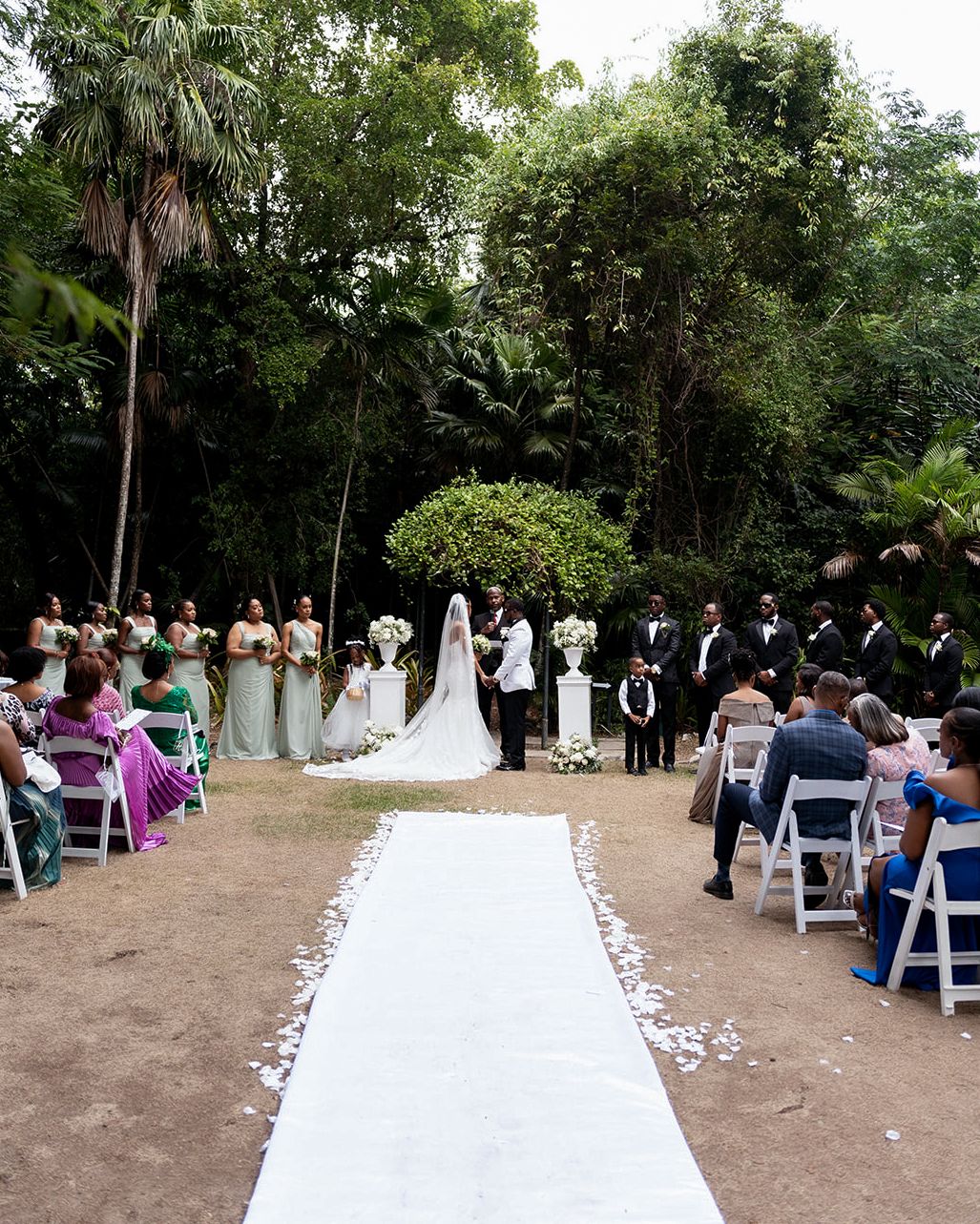 Outdoor wedding ceremony with bride and groom at the altar, bridesmaids in sage green, white aisle runner, and lush tropical greenery.