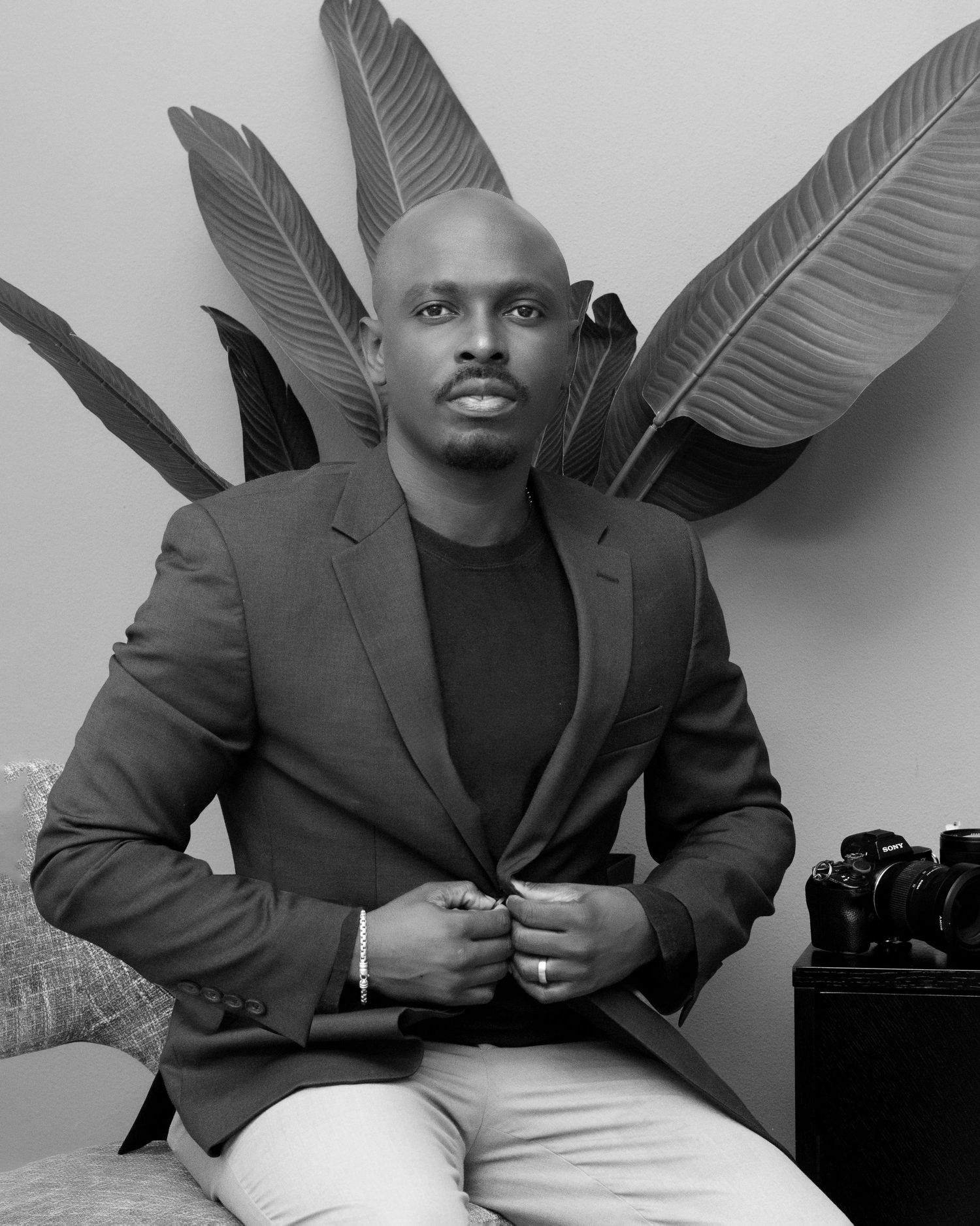 Black and white portrait of a well-dressed man in a blazer seated confidently with tropical leaves in the background.