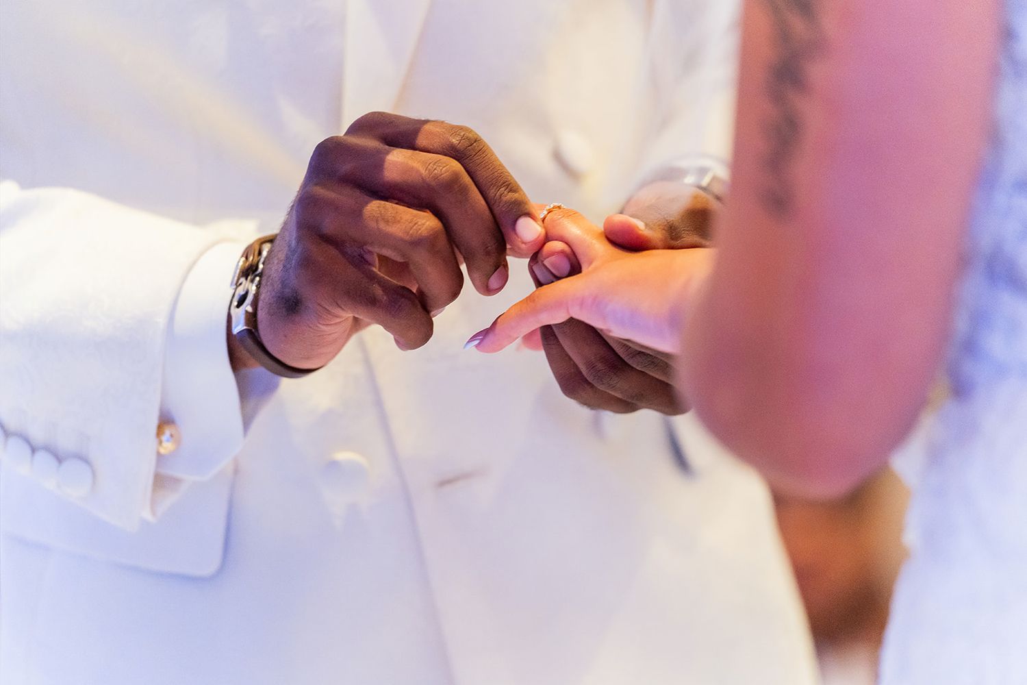 Groom in white suit placing a wedding ring on bride's finger during a ceremony.