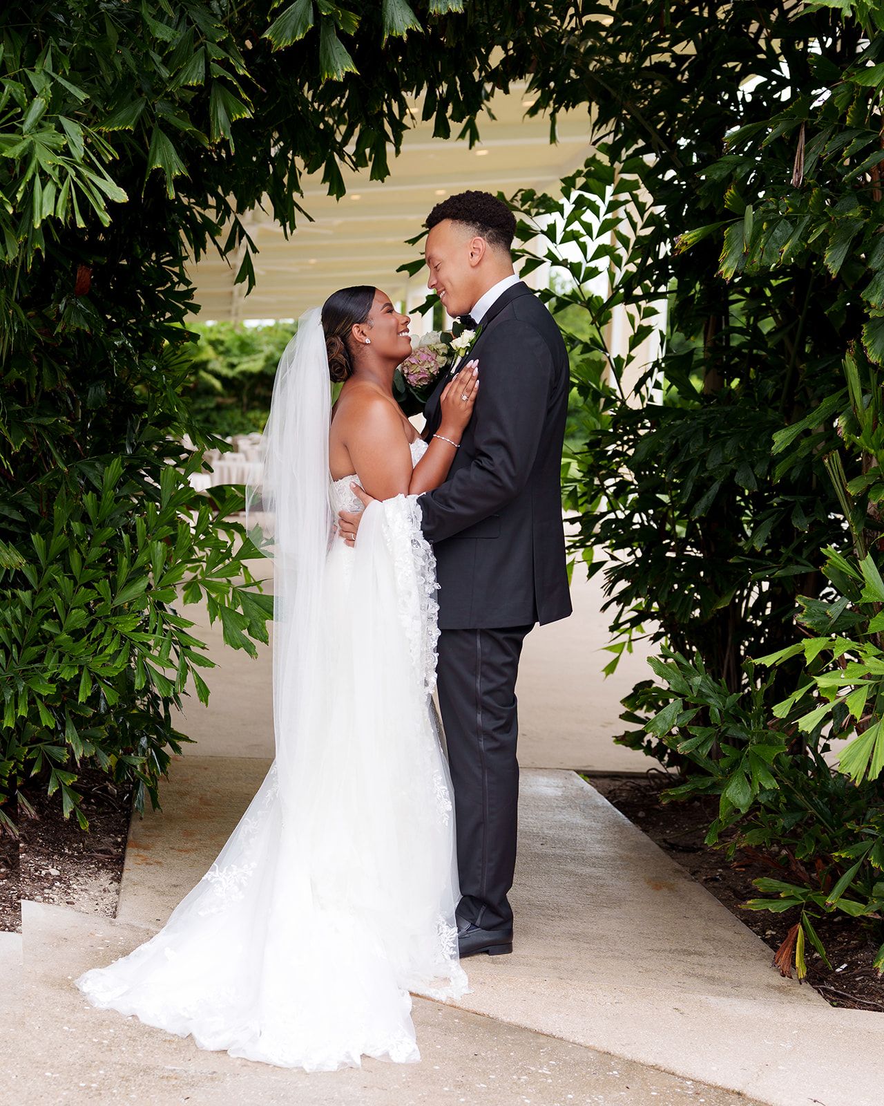 Bride in lace mermaid gown and veil gazes at groom in black suit under lush green garden archway.