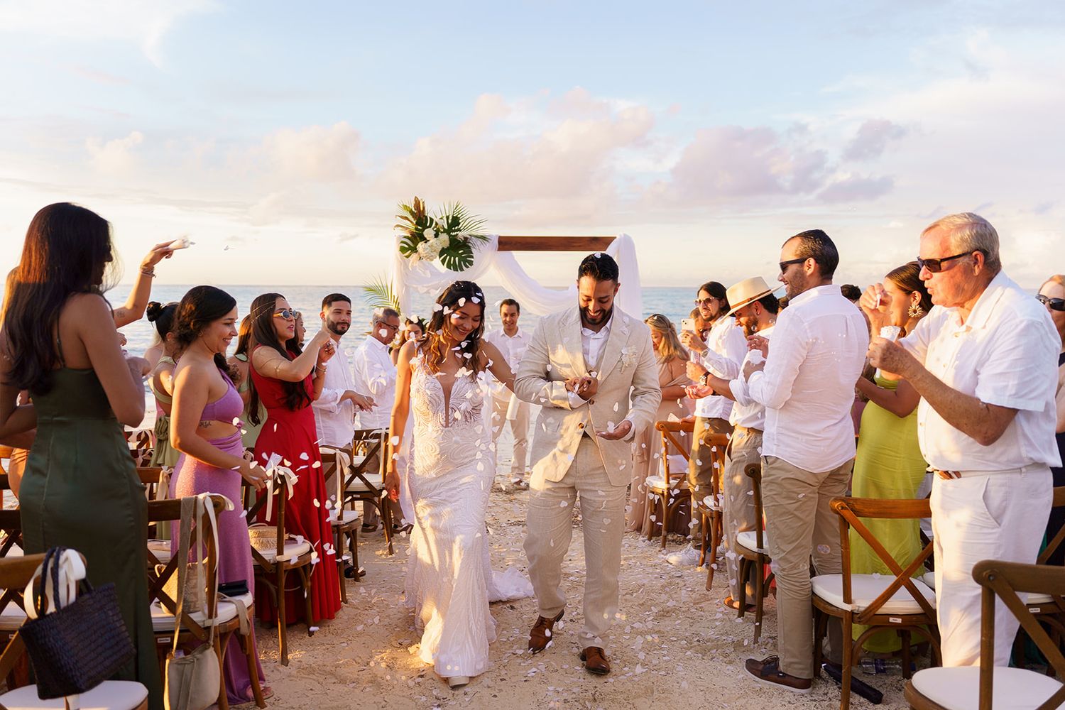 Couple walks down the aisle at an outdoor beach wedding ceremony surrounded by cheering guests at sunset.
