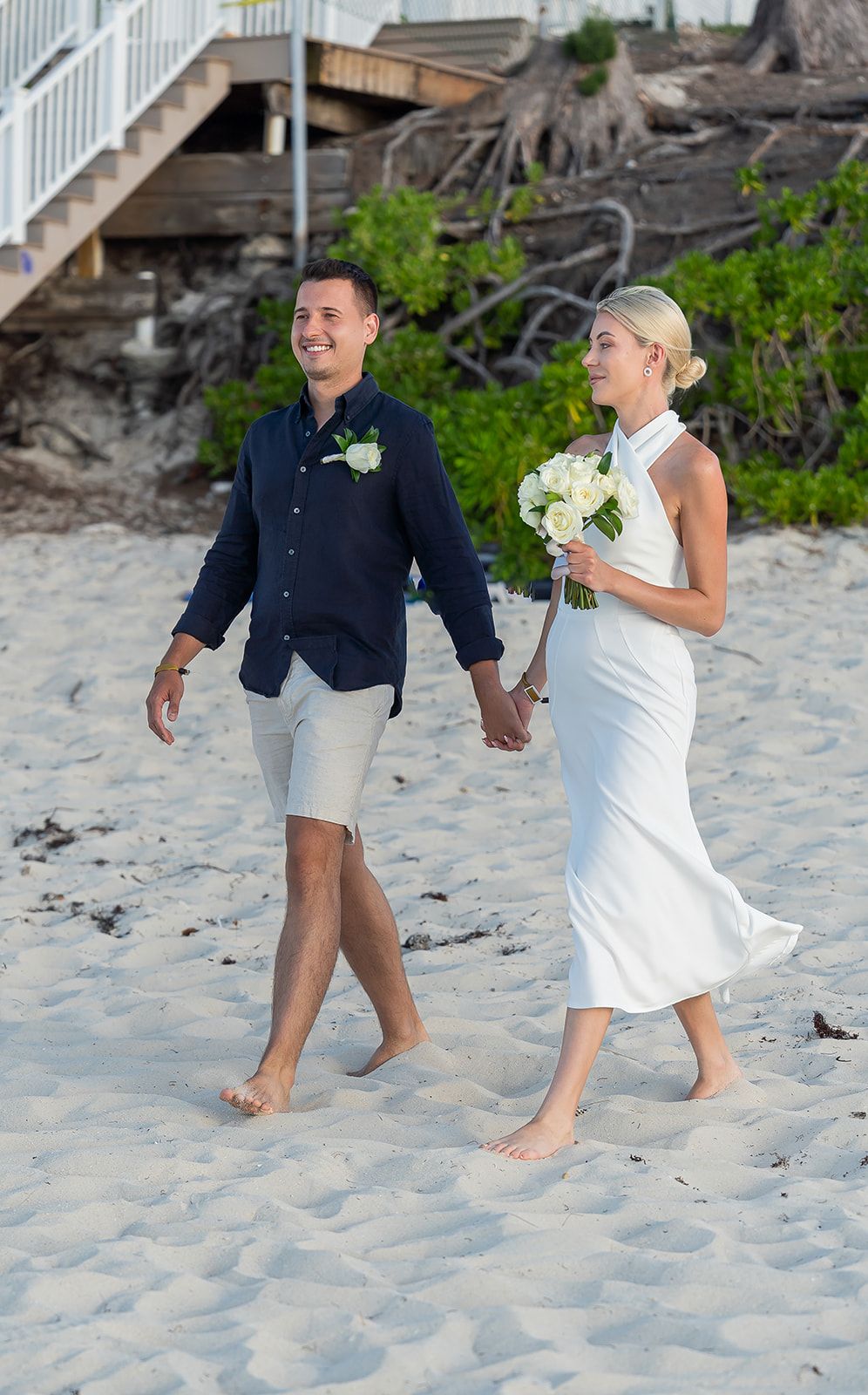 Couple in wedding attire walking barefoot on white sand beach, bride in white halter dress holding bouquet, groom in navy shirt.