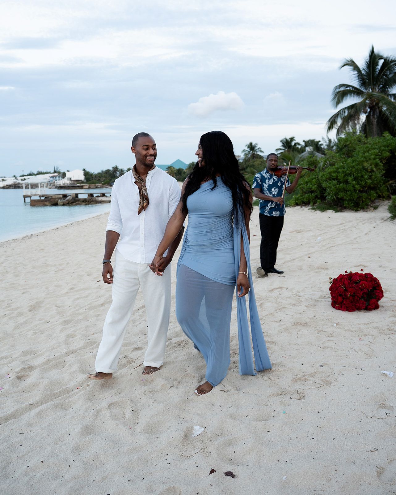 Couple holding hands on a tropical beach, man in white linen outfit, woman in light blue dress, red roses nearby.