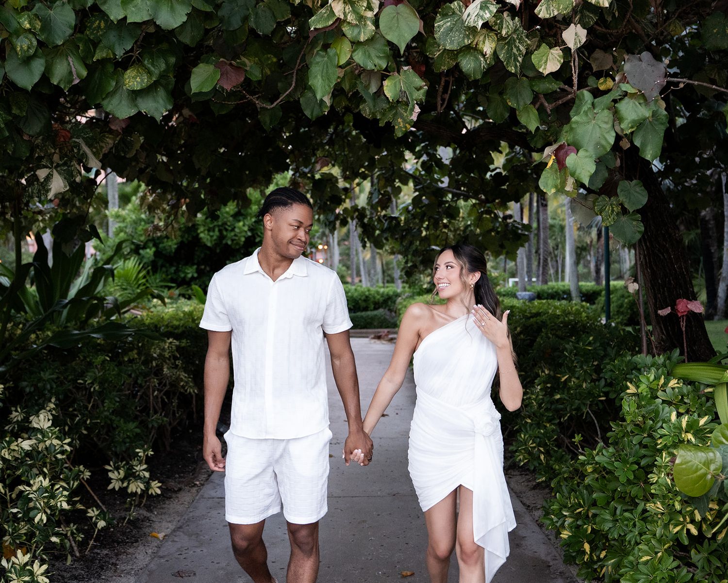 Couple in matching white outfits holding hands and walking along a lush garden path lined with green trees.