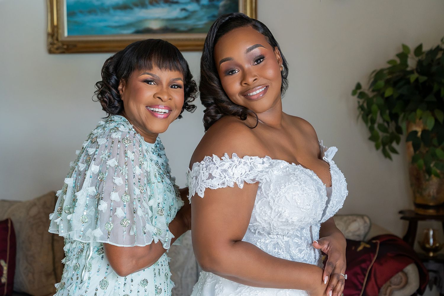 A bride in a white off-shoulder gown smiles with an older woman in a mint lace dress indoors.