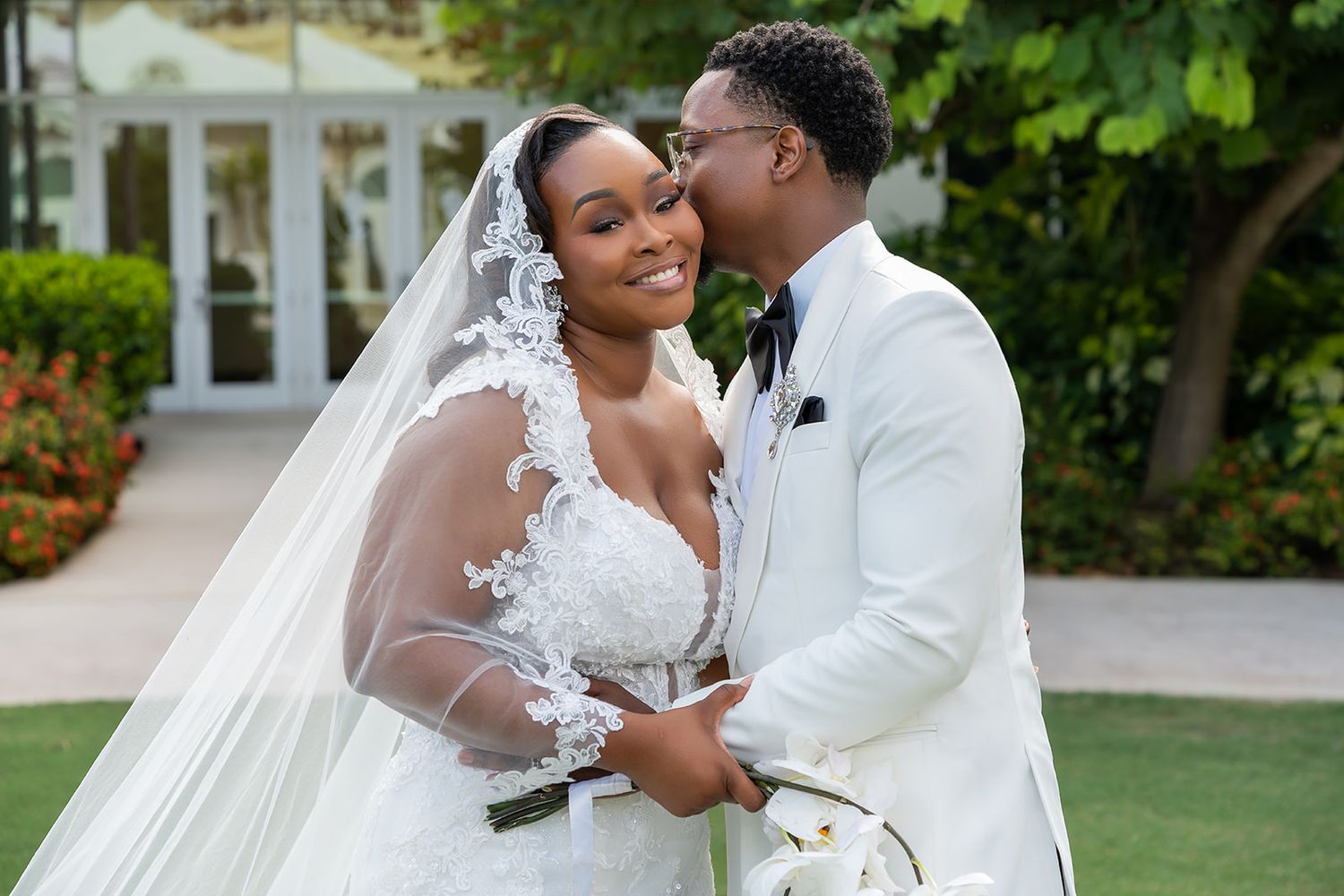 A Black couple in wedding attire embraces outdoors, bride in lace gown with veil, groom in white tuxedo.