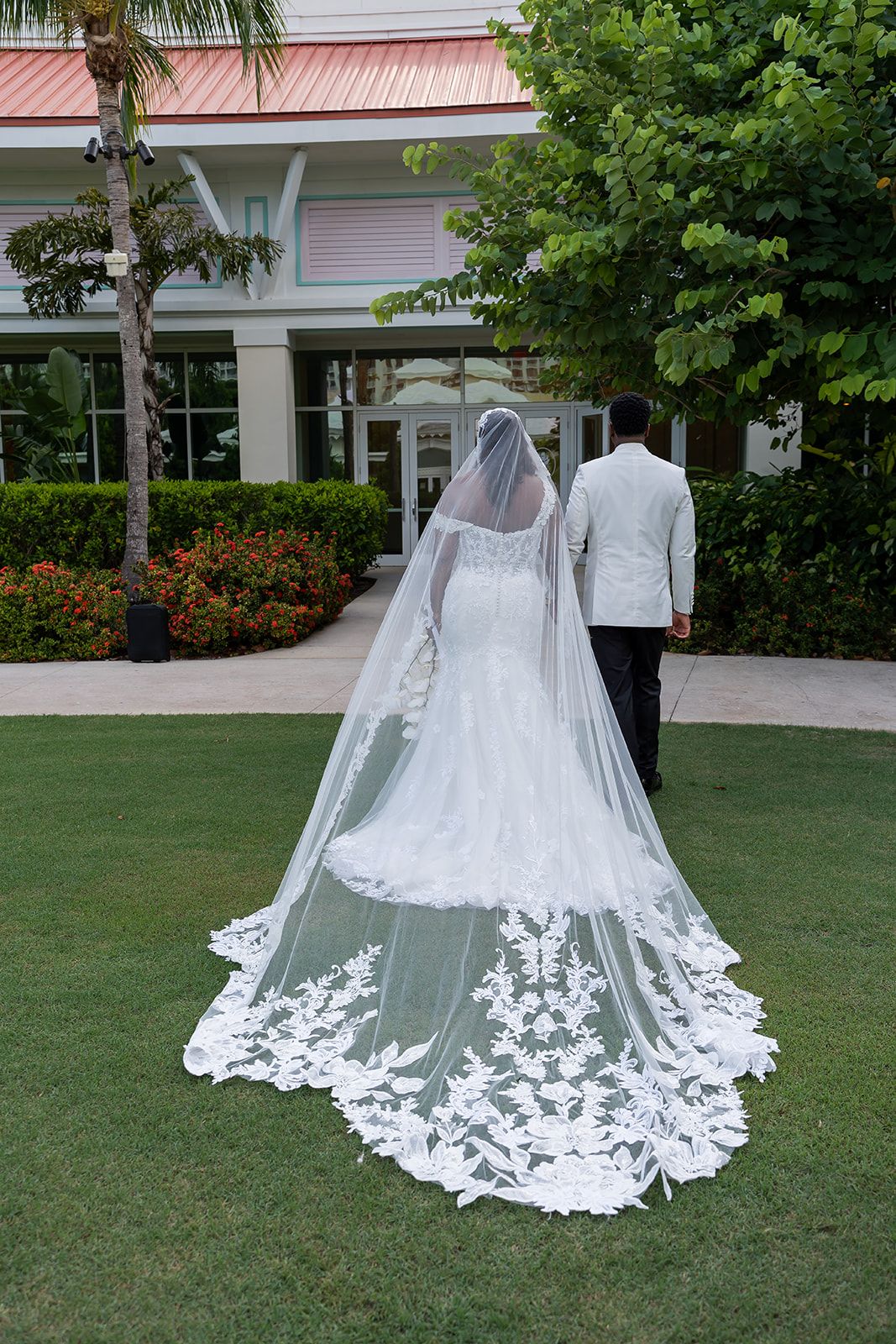 Bride in lace wedding gown with cathedral veil walks with groom in white suit on green lawn near tropical venue.