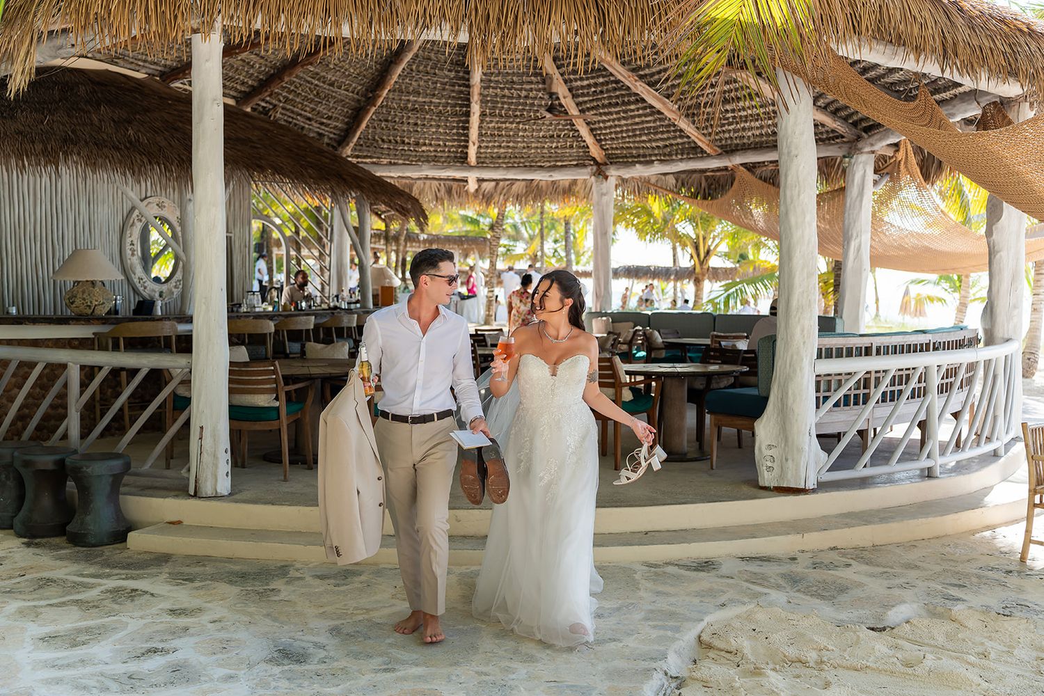 Bride and groom walking hand in hand under a tropical thatched-roof beach bar at a resort wedding.