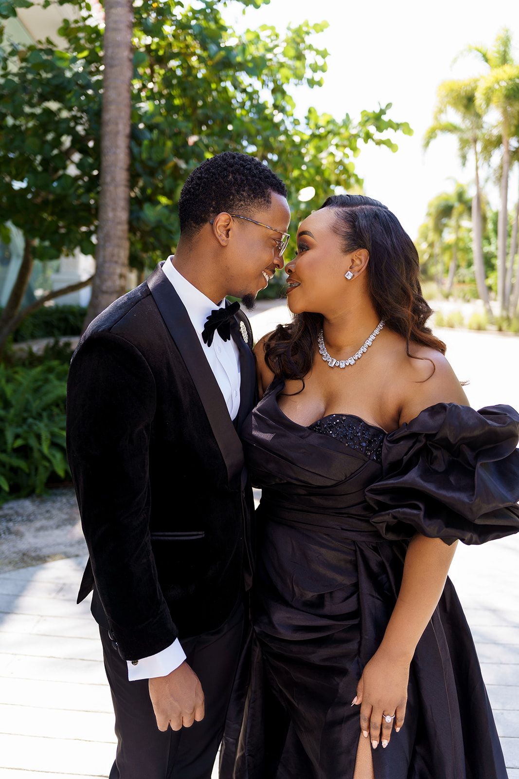 Elegant couple in black formal attire posing outdoors, man in tuxedo and woman in off-shoulder gown with diamond jewelry.
