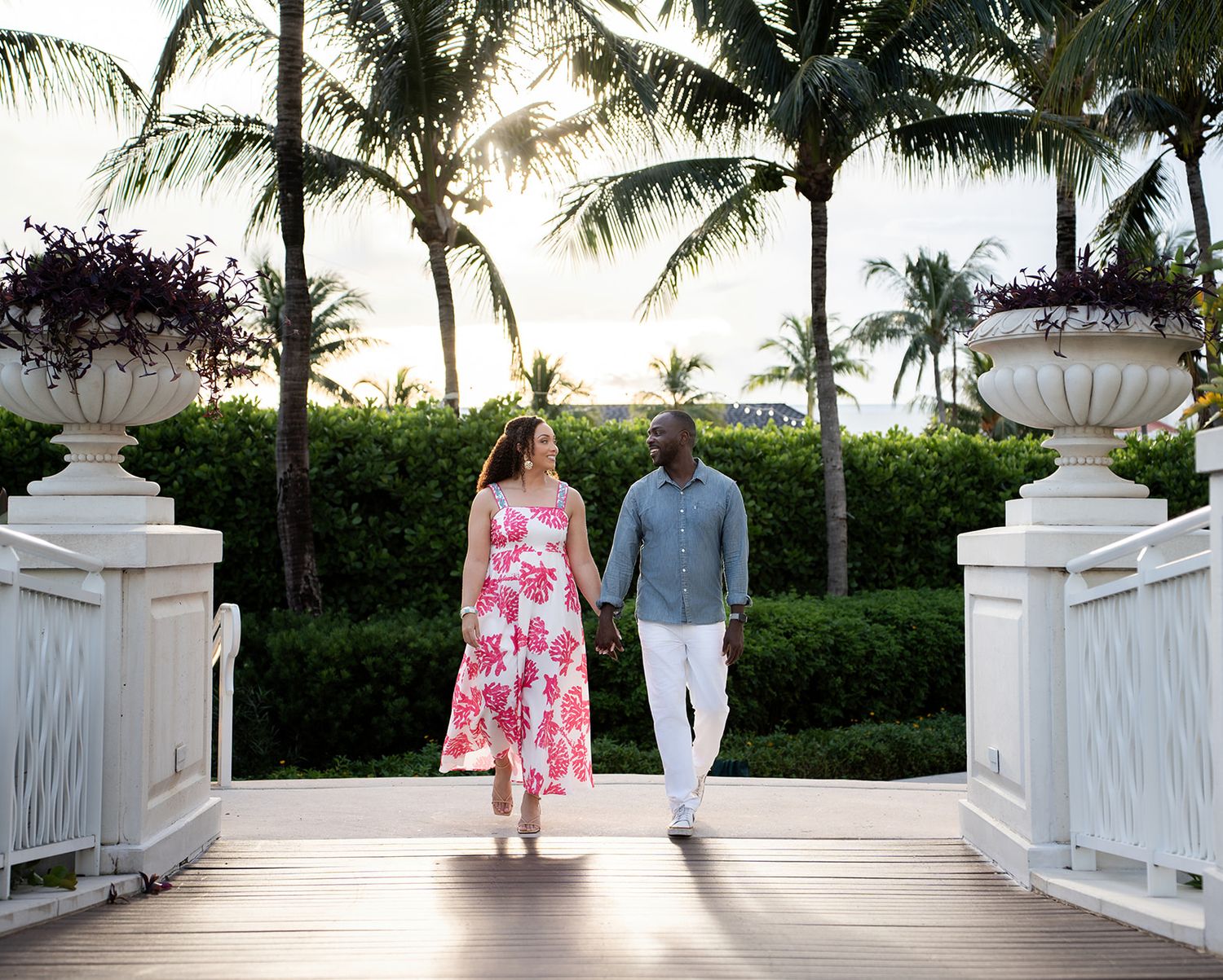 Couple holding hands walking on a wooden bridge surrounded by palm trees and tropical greenery at sunset.