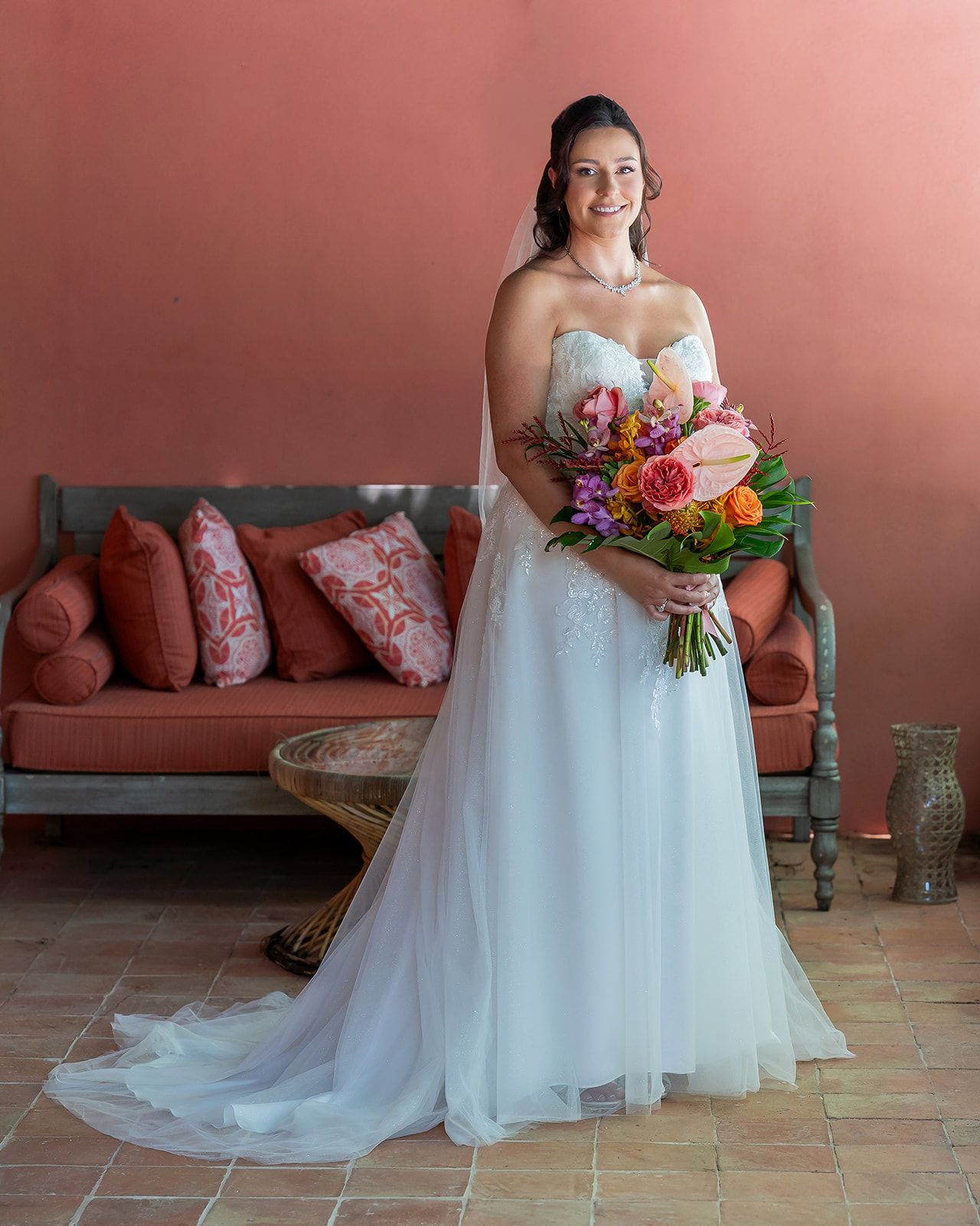 Bride in white strapless gown holding colorful bouquet stands before coral wall with ornate sofa.
