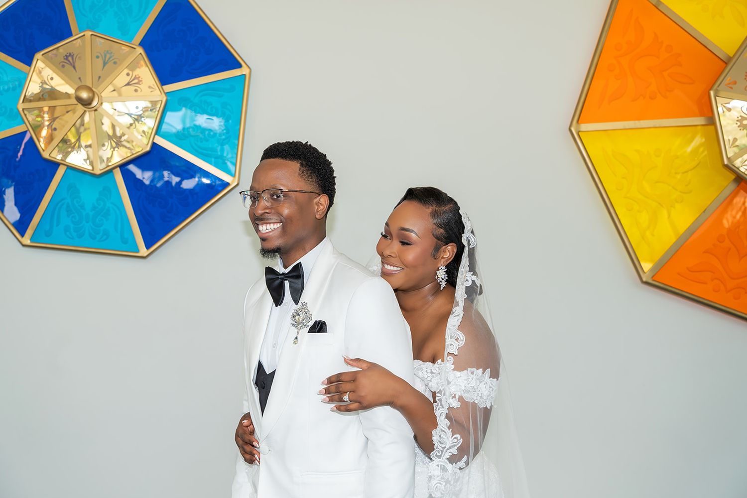 Happy couple in wedding attire smiling together, groom in white tuxedo and bride in lace gown, colorful stained glass decor.