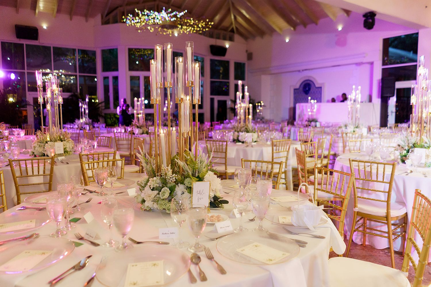 Luxurious wedding reception table set with tall candelabras, white floral arrangements, gold Chiavari chairs, and purple uplighting.