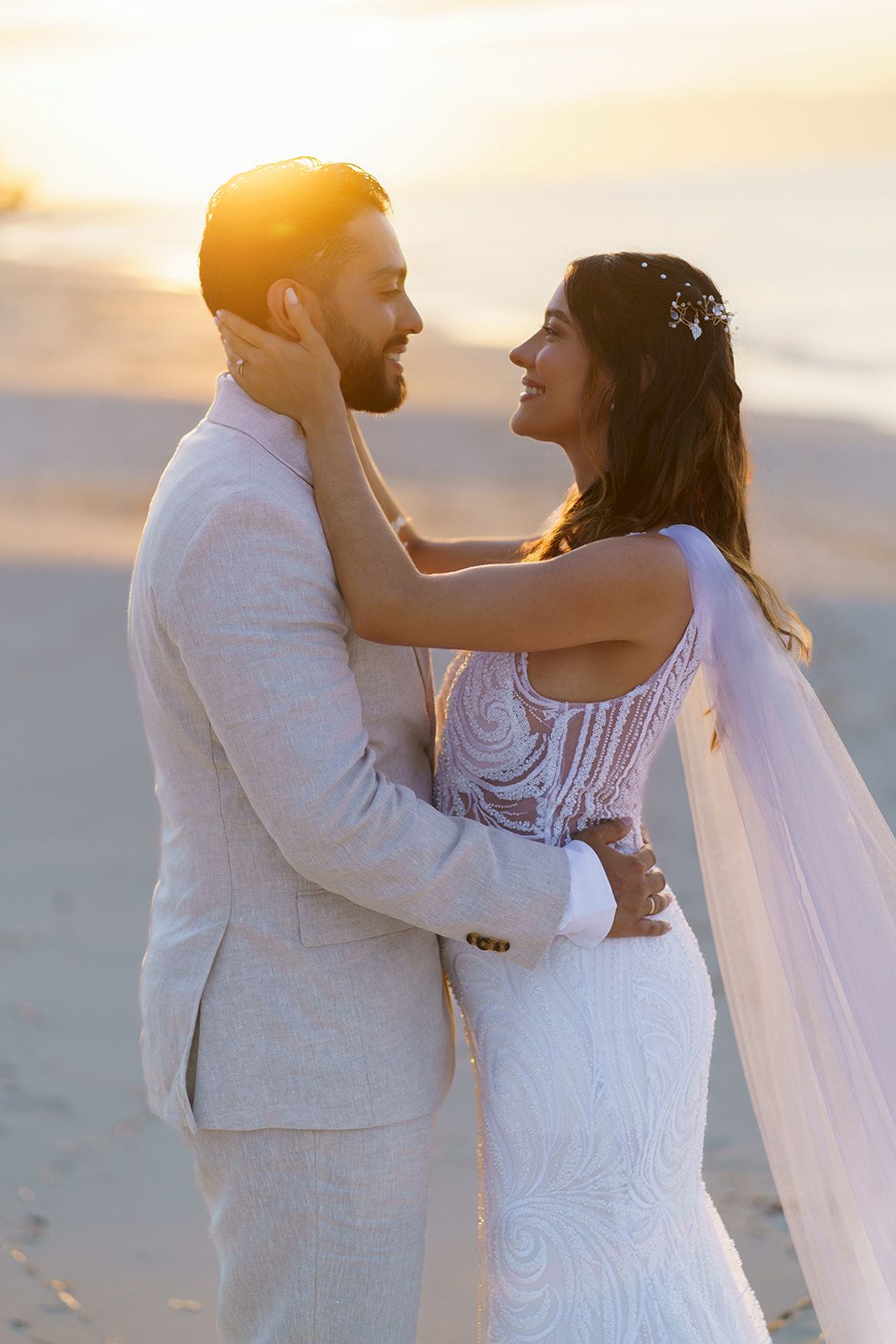 Bride and groom embrace on a sandy beach at golden sunset, she wears a beaded gown with veil, he wears a light suit.