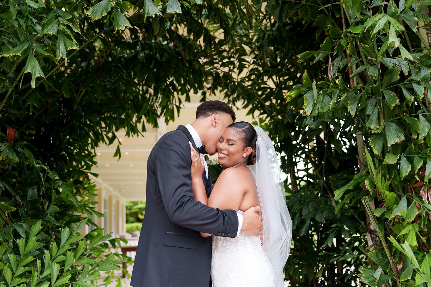 Bride and groom share a romantic kiss under a lush green leafy arch on their wedding day.