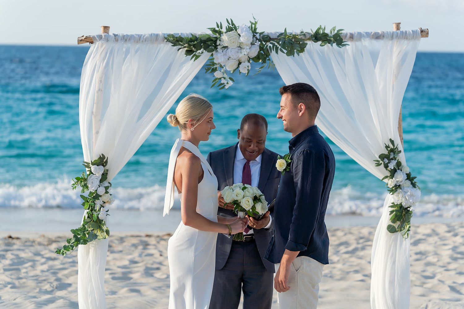 Couple exchanging vows at a beach wedding under a white floral arch with ocean backdrop.