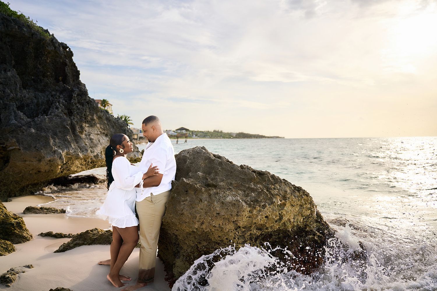 Couple embracing romantically on a tropical beach at sunset beside large rocks with ocean waves.