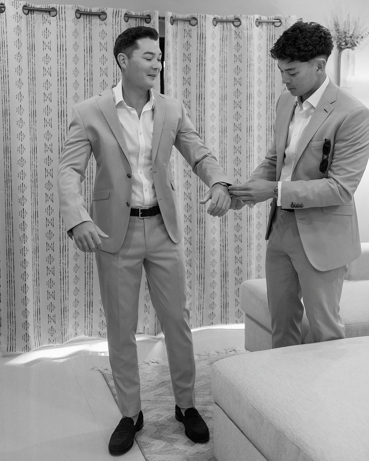 Two men in matching light suits adjusting cufflinks in a black and white photo at a formal event.