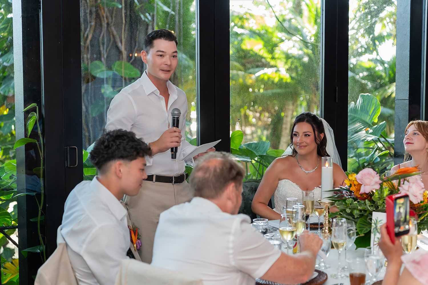 Groomsman delivers heartfelt speech at wedding reception as smiling bride and groom listen at floral decorated table.