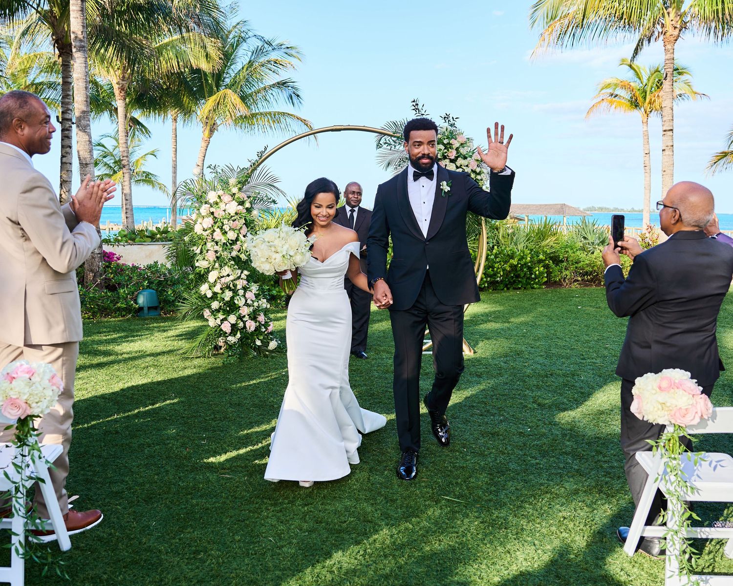 Wedding party dances on a green lawn surrounded by palm trees at a tropical beachside venue on a sunny day.
