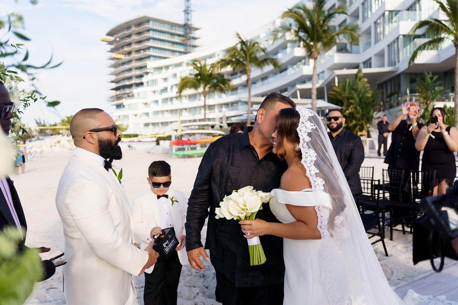 Bride and groom sharing a kiss at a beachfront wedding ceremony with guests and palm trees in the background.