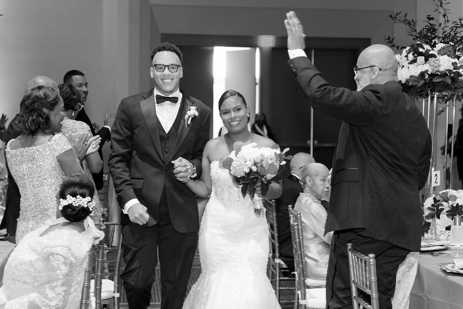 Bride and groom walk down aisle holding hands at elegant wedding reception in black and white photo.