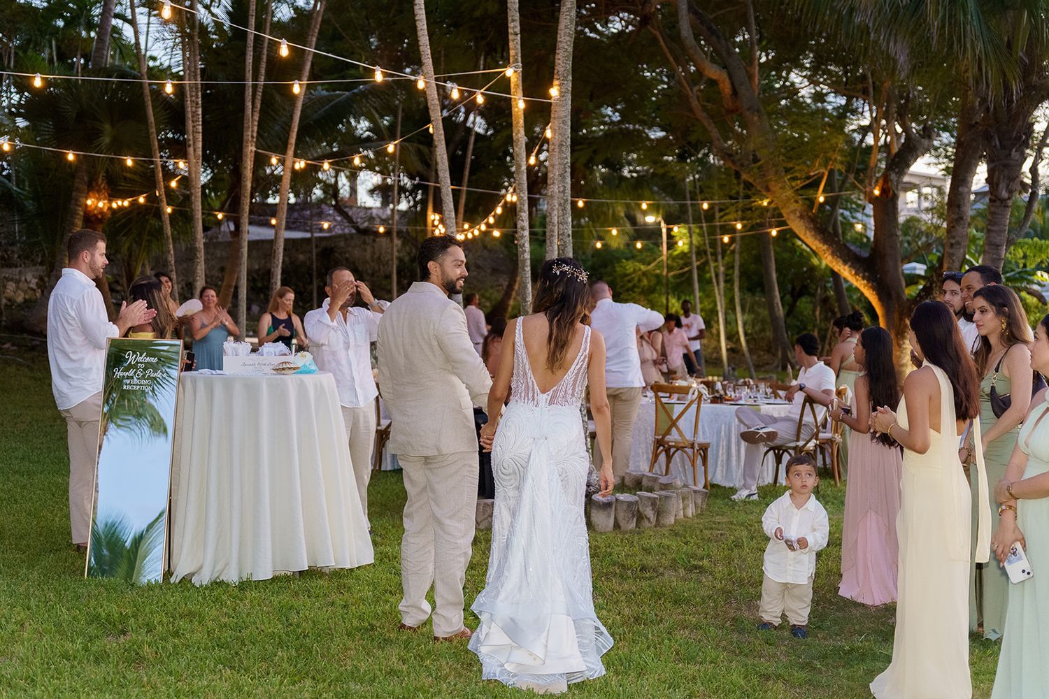 Outdoor wedding reception under string lights with bride and groom walking among guests on tropical garden lawn.