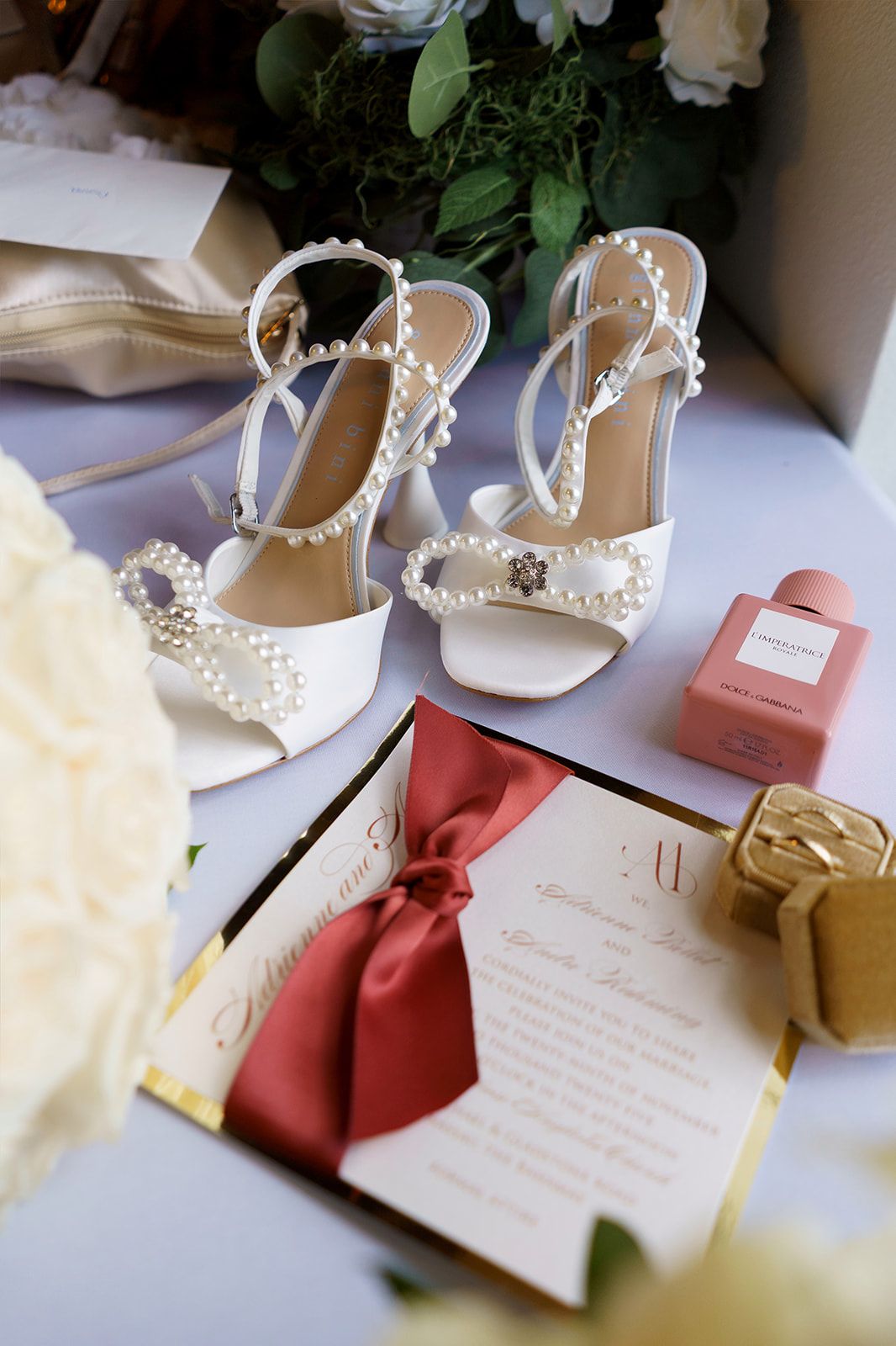 White pearl-embellished bridal heels displayed with wedding invitation, pink perfume, and white roses.