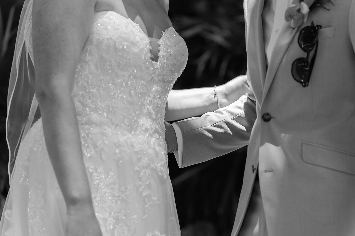 Black and white photo of bride and groom holding hands during wedding ceremony, bride in lace gown with veil.