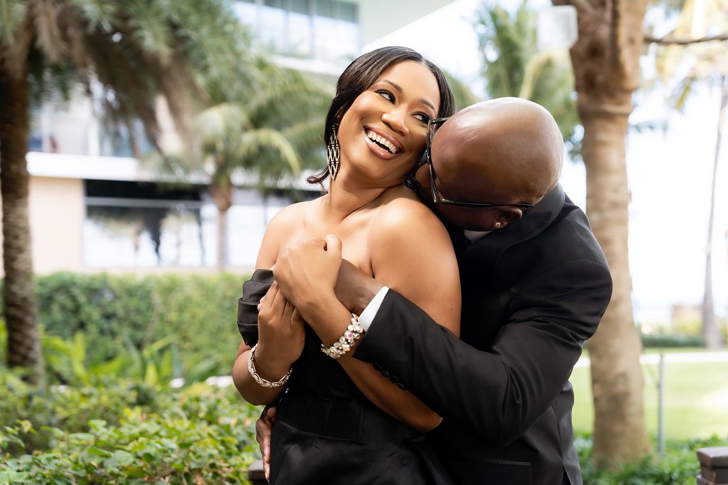 A joyful couple embraces outdoors, woman laughing in black strapless dress, man hugging her from behind near palm trees.