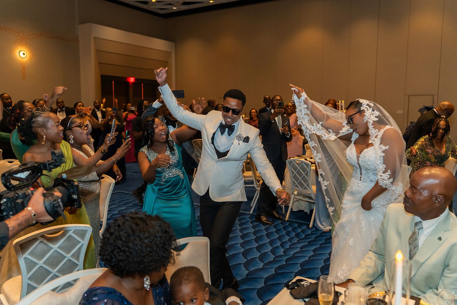 Joyful newlyweds make grand entrance at wedding reception, groom in white tuxedo, bride in veil, guests celebrating.