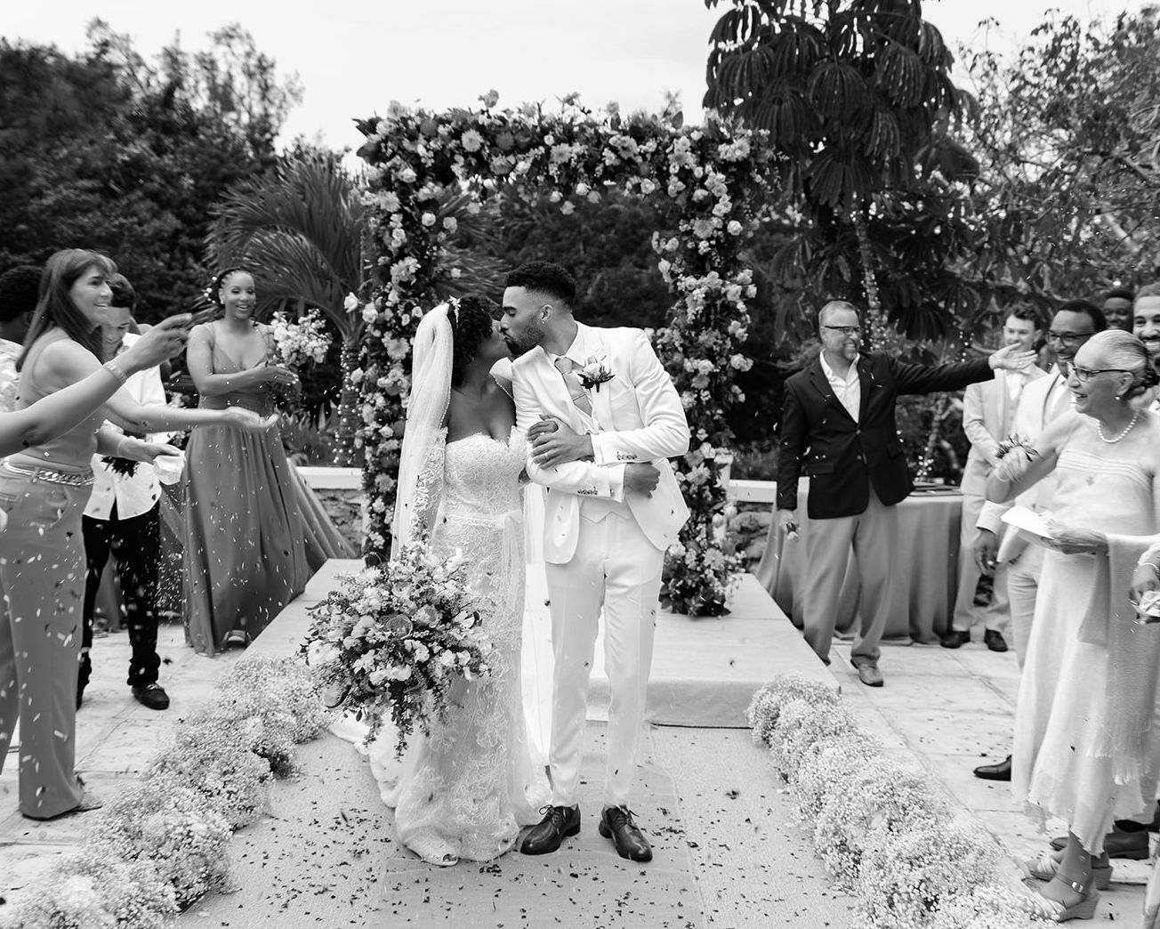 Black and white photo of a couple sharing a first kiss at their outdoor wedding ceremony with floral arch.