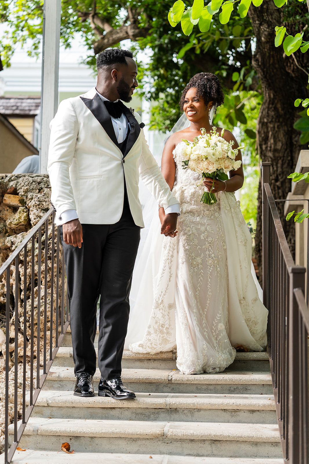 Bride and groom holding hands on outdoor stairs, bride in lace gown, groom in white tuxedo jacket.