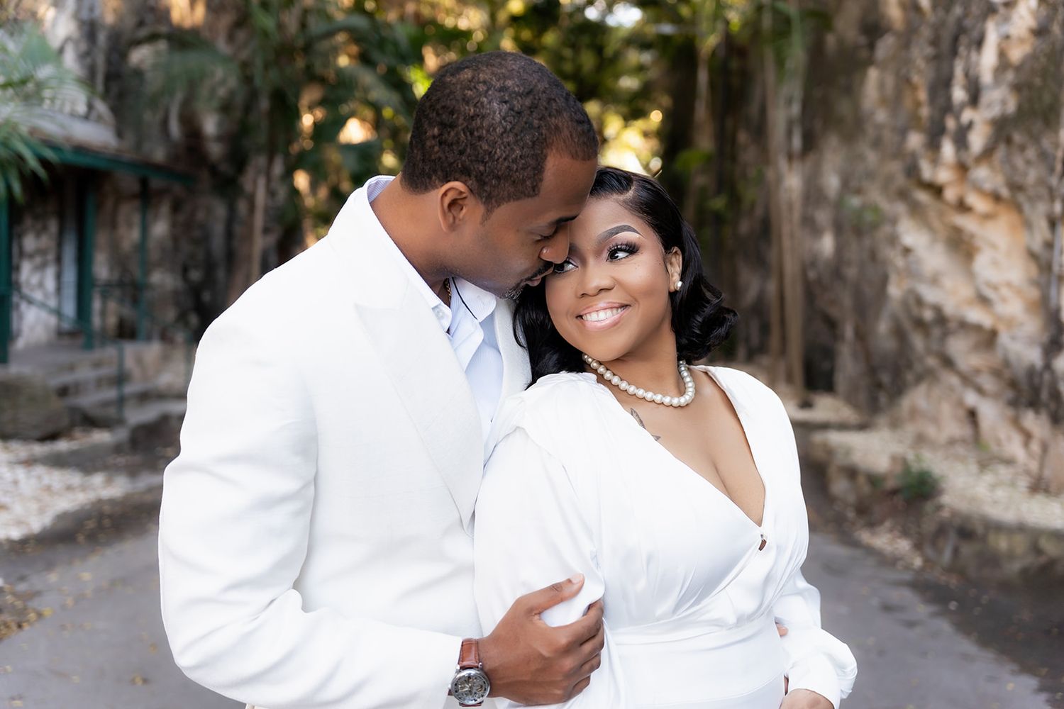 A couple in white attire shares a romantic embrace outdoors, with the man tenderly nuzzling the smiling woman wearing pearls.