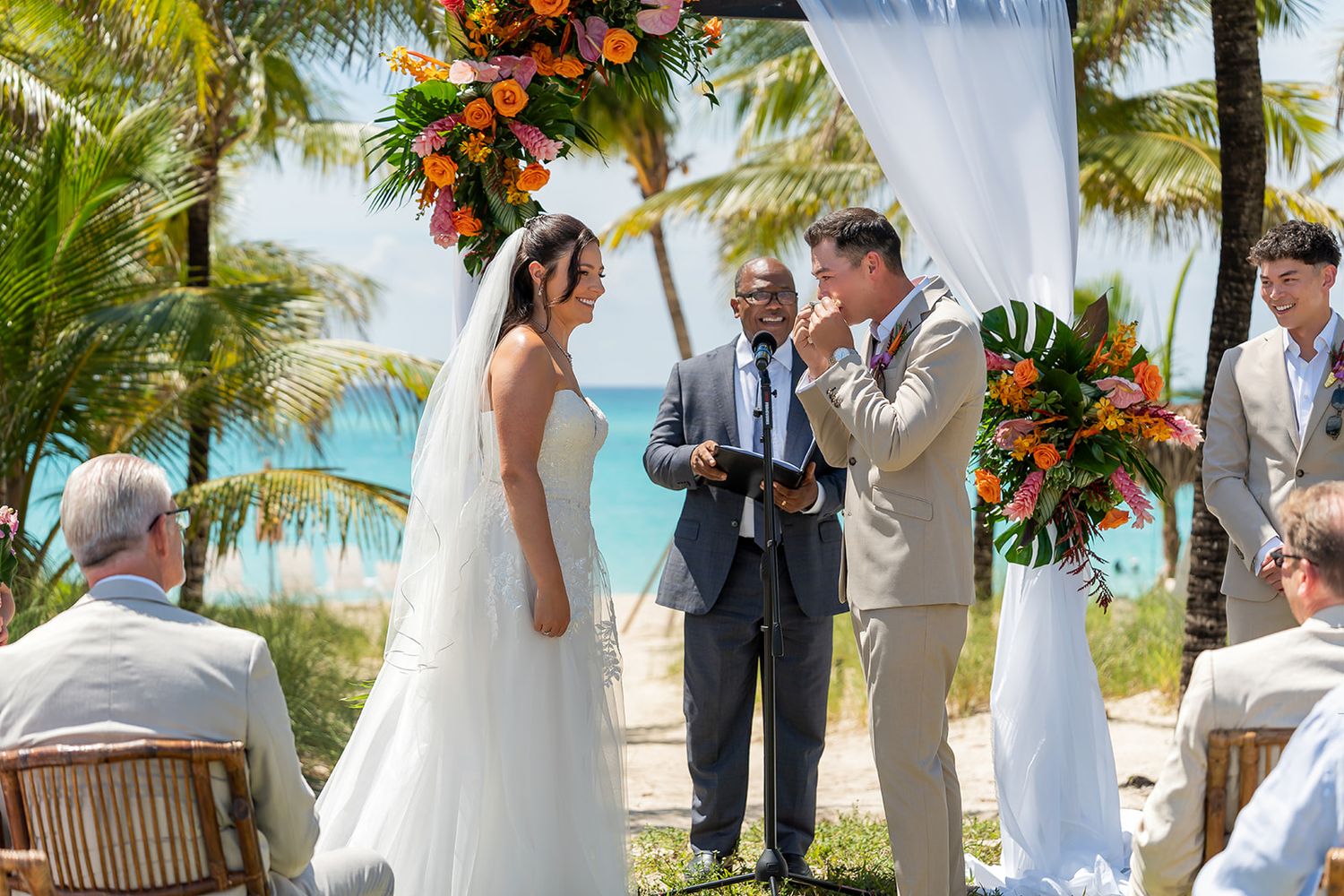Couple exchanging vows at a tropical beach wedding ceremony with palm trees and vibrant orange floral arch.