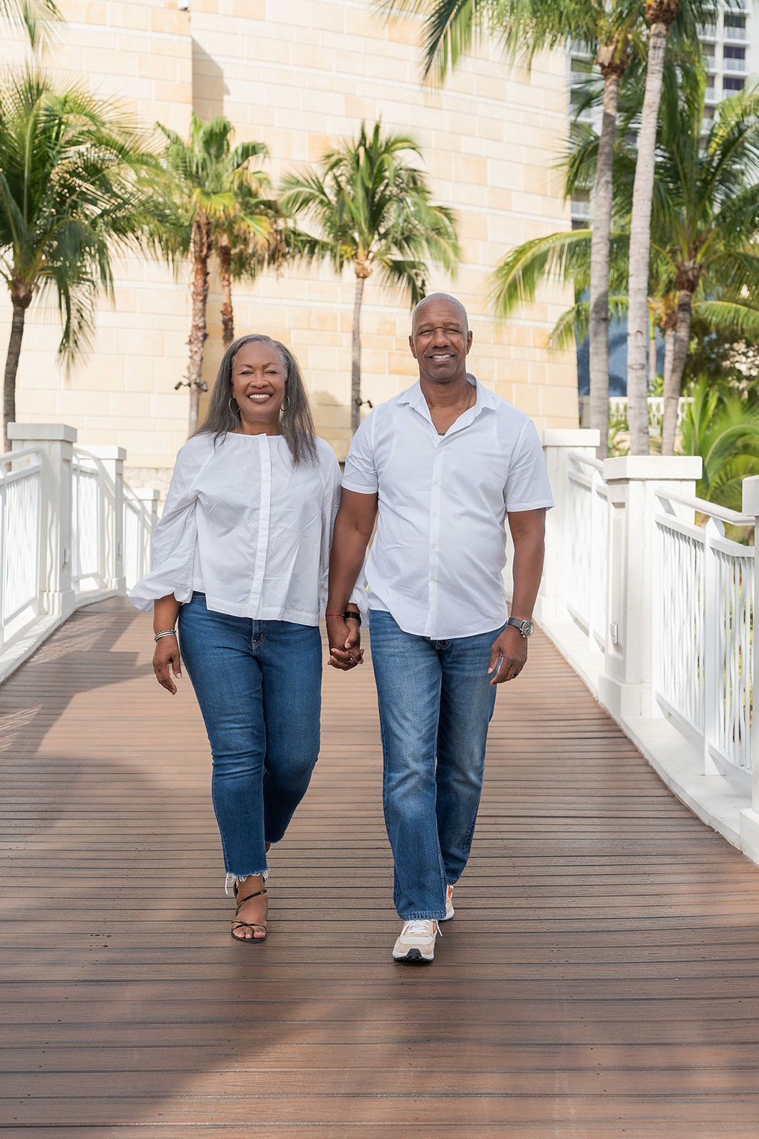 Happy senior couple holding hands and walking on a wooden bridge surrounded by palm trees in a tropical setting.