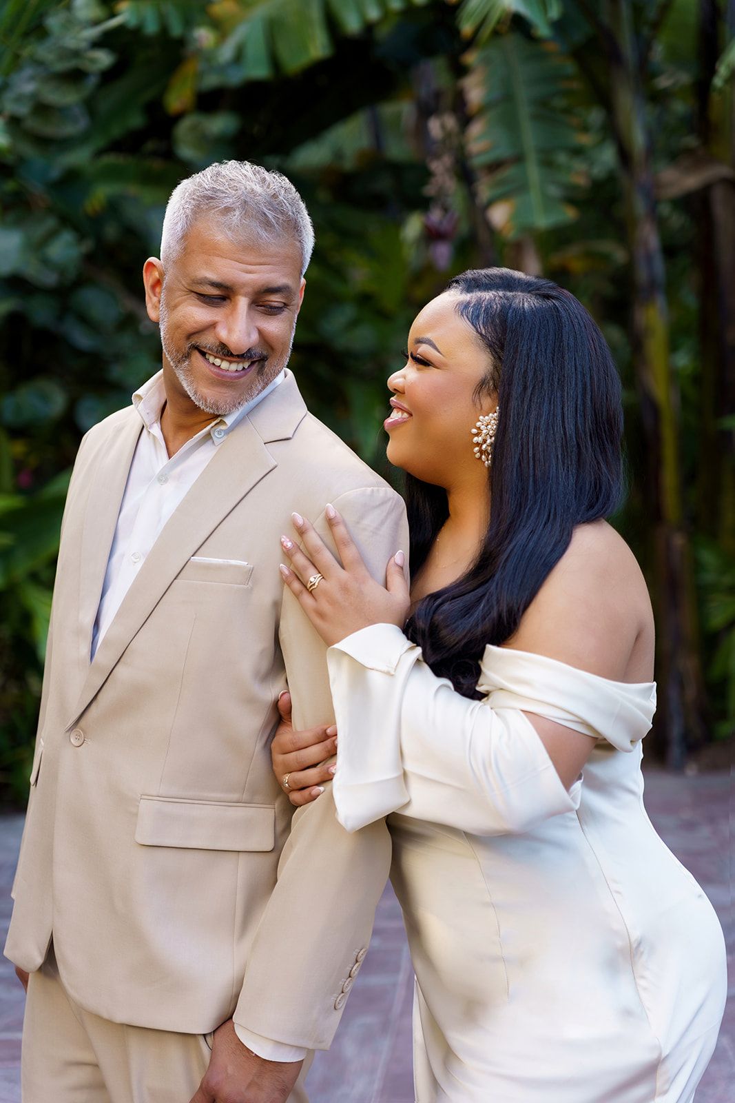 Elegant couple in formal attire smiling together outdoors, woman in white off-shoulder dress, man in beige suit.