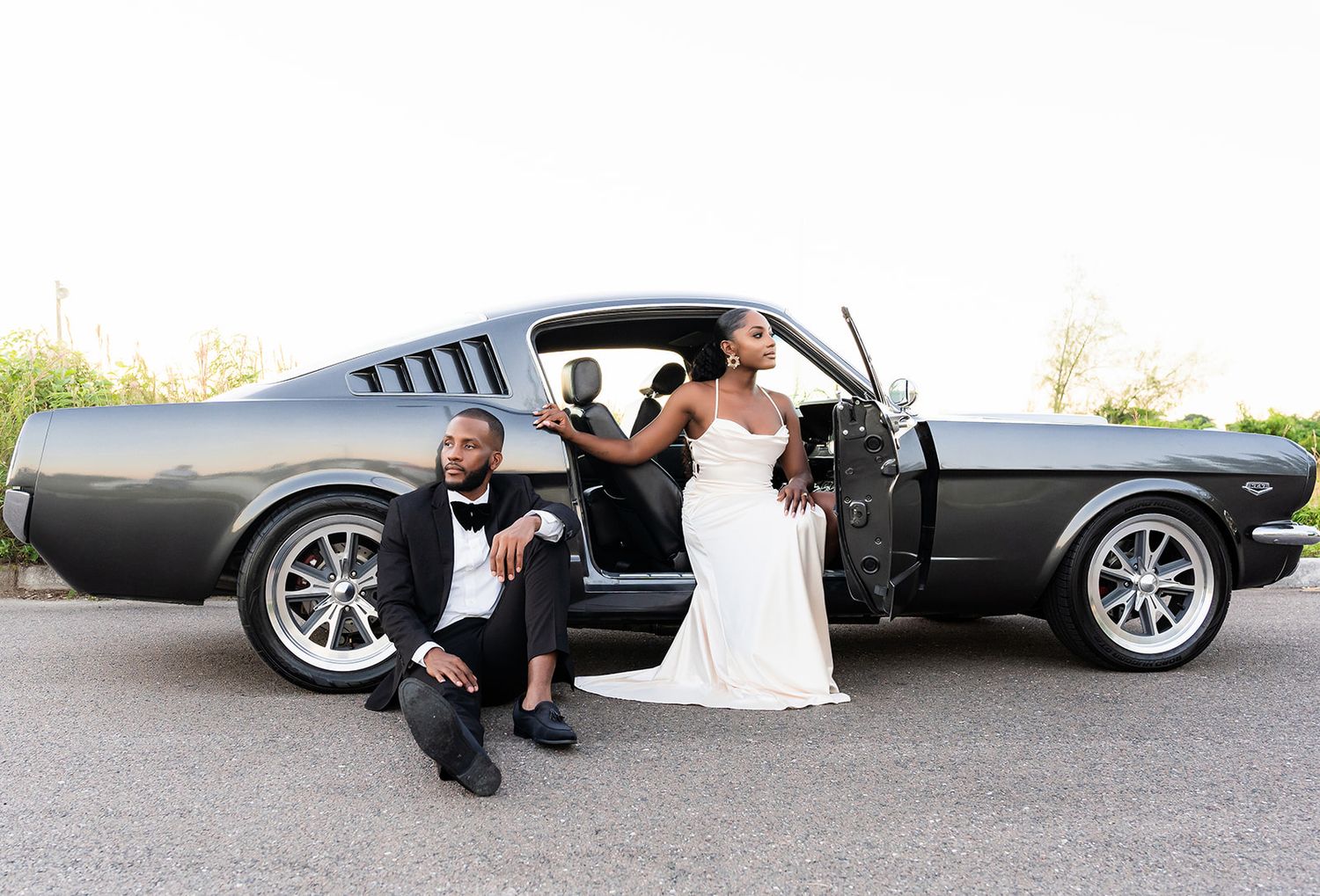 Bride and groom posing with a classic black Ford Mustang on their wedding day.