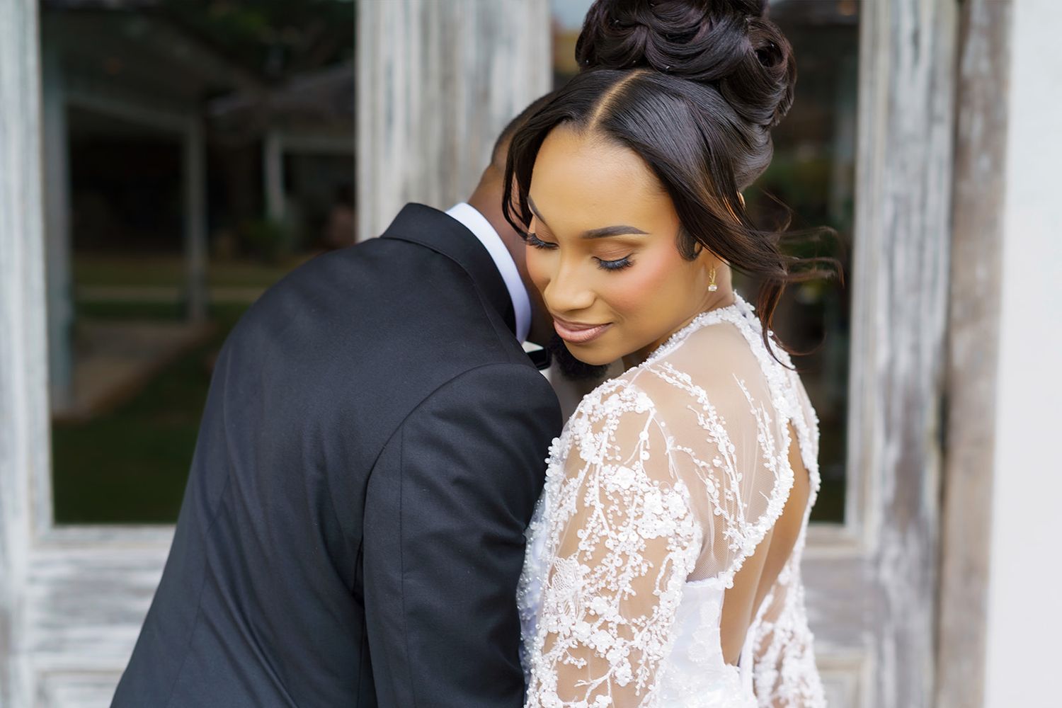 Bride in lace long-sleeve wedding gown embraces groom in black suit during romantic outdoor portrait.