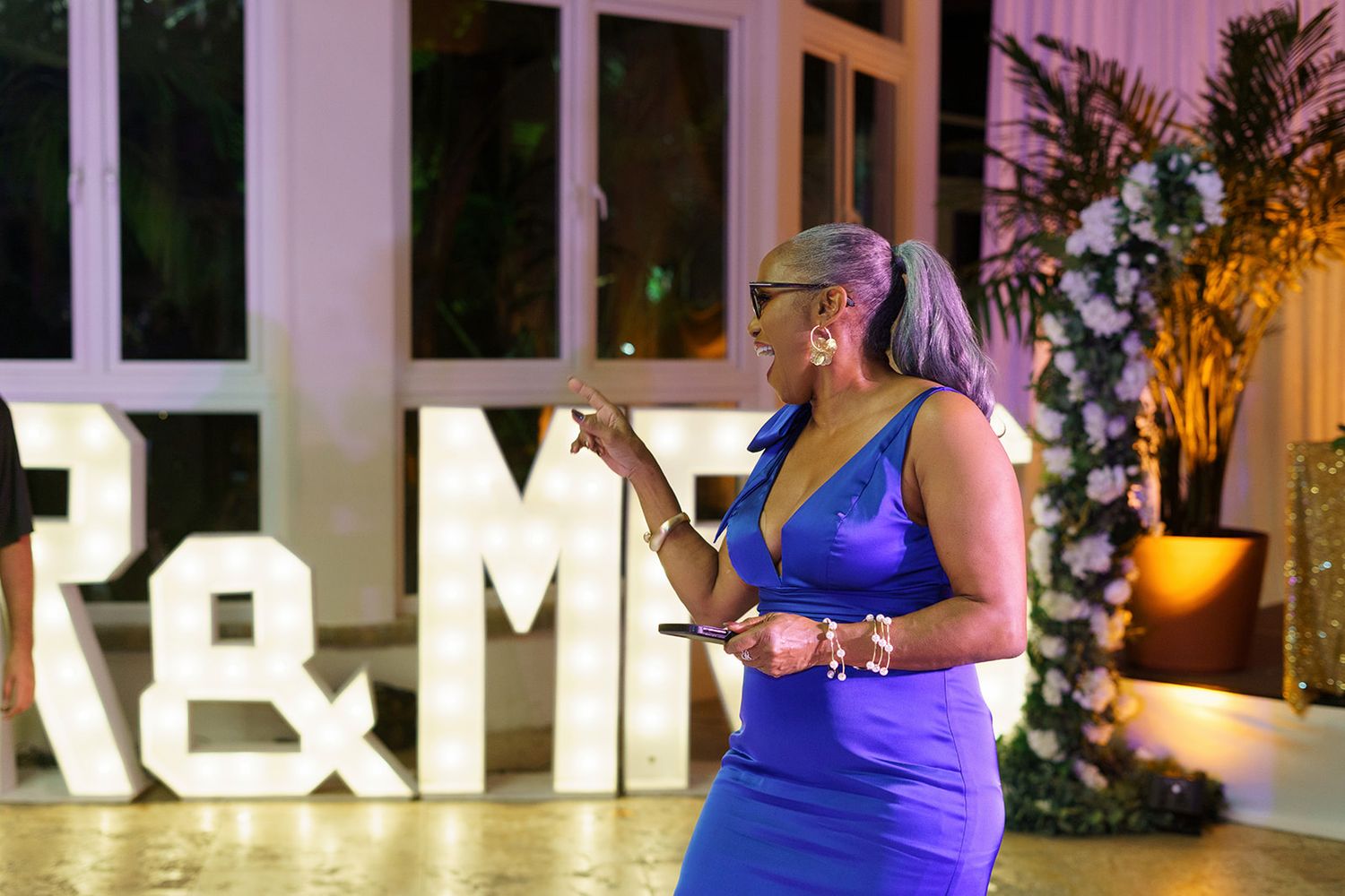 A woman in a blue dress speaks at a wedding reception beside illuminated R&M letter signs.