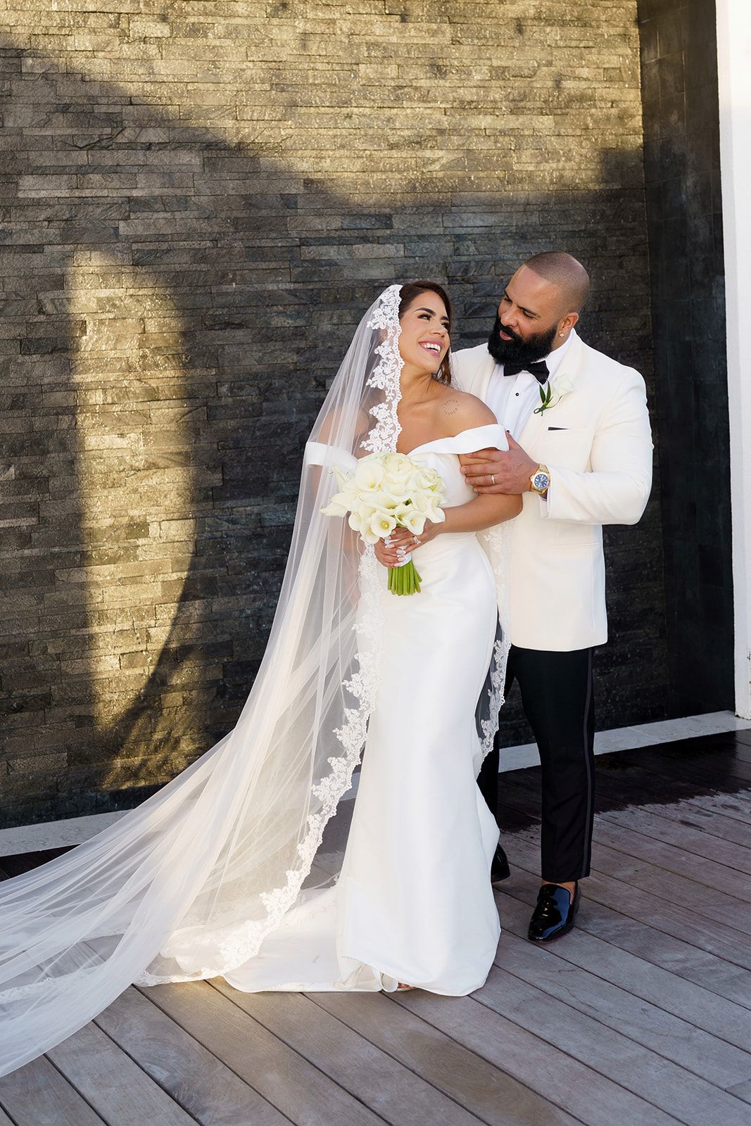 Bride in white off-shoulder gown with lace veil holds bouquet as groom in white tuxedo embraces her outdoors.
